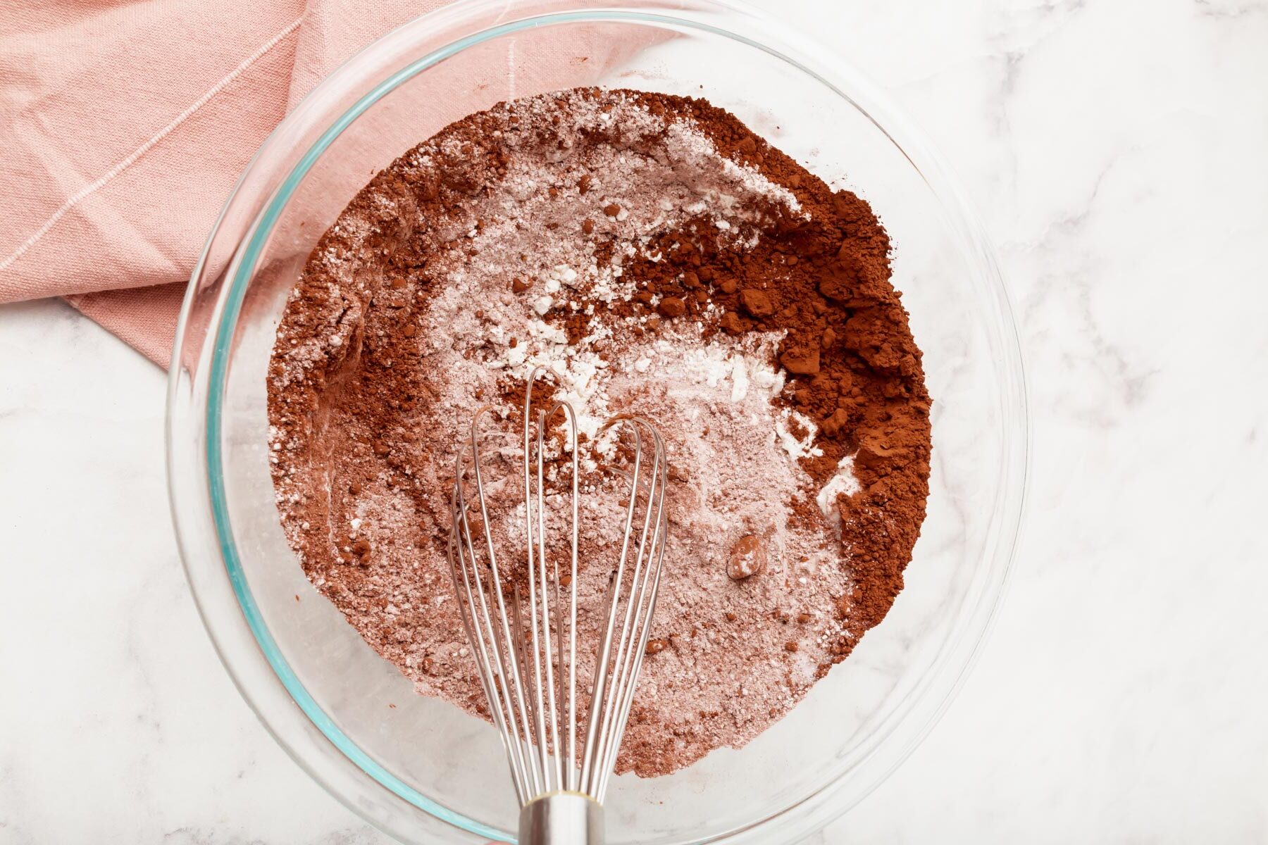 A glass bowl filled with a mixture of cocoa powder and flour on a marble surface, with a metal whisk resting inside. A pink cloth is partially visible in the corner.