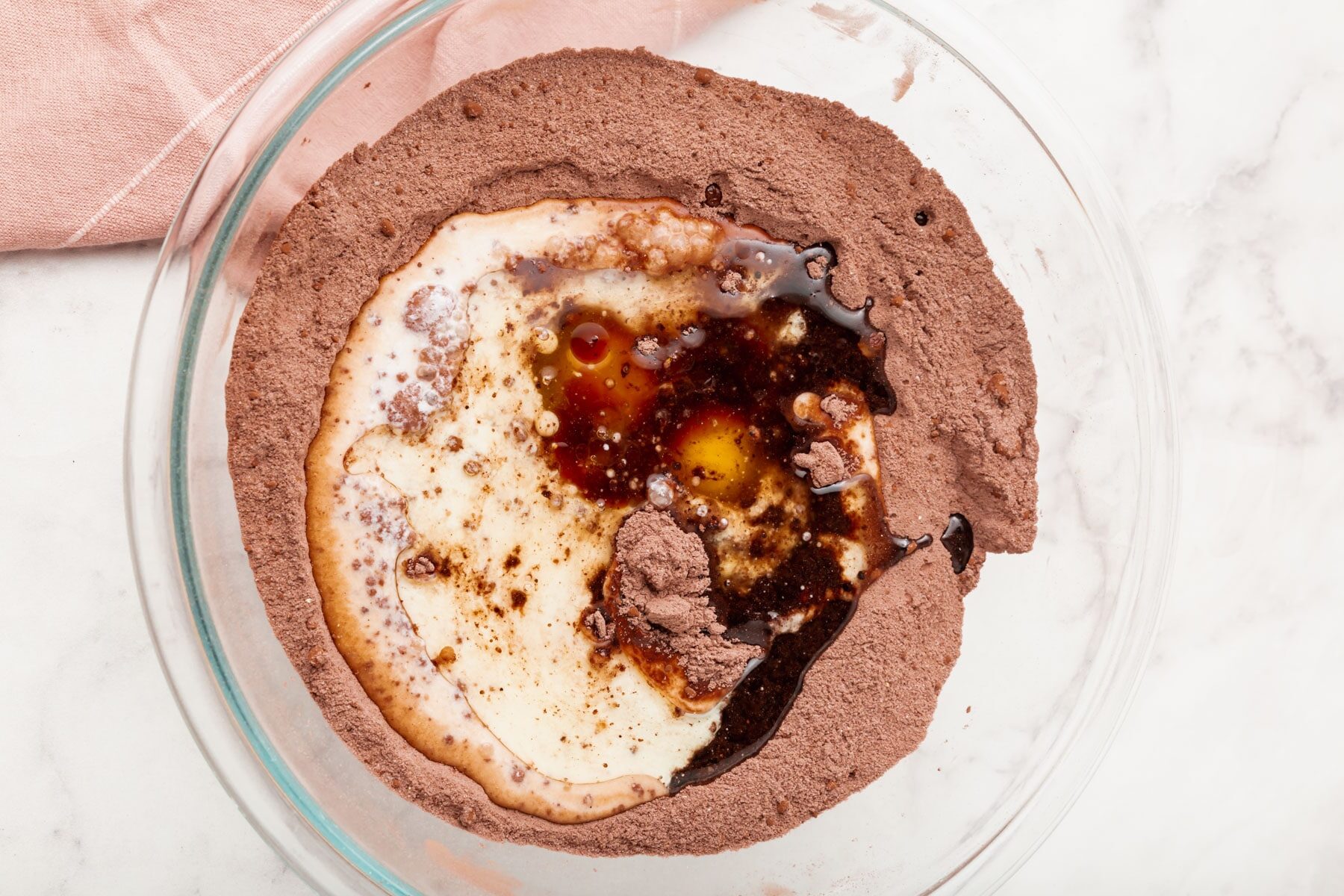 A glass bowl filled with dry chocolate cake mix, eggs, milk, and vanilla extract, about to be mixed together on a light marble surface with a pink cloth nearby.