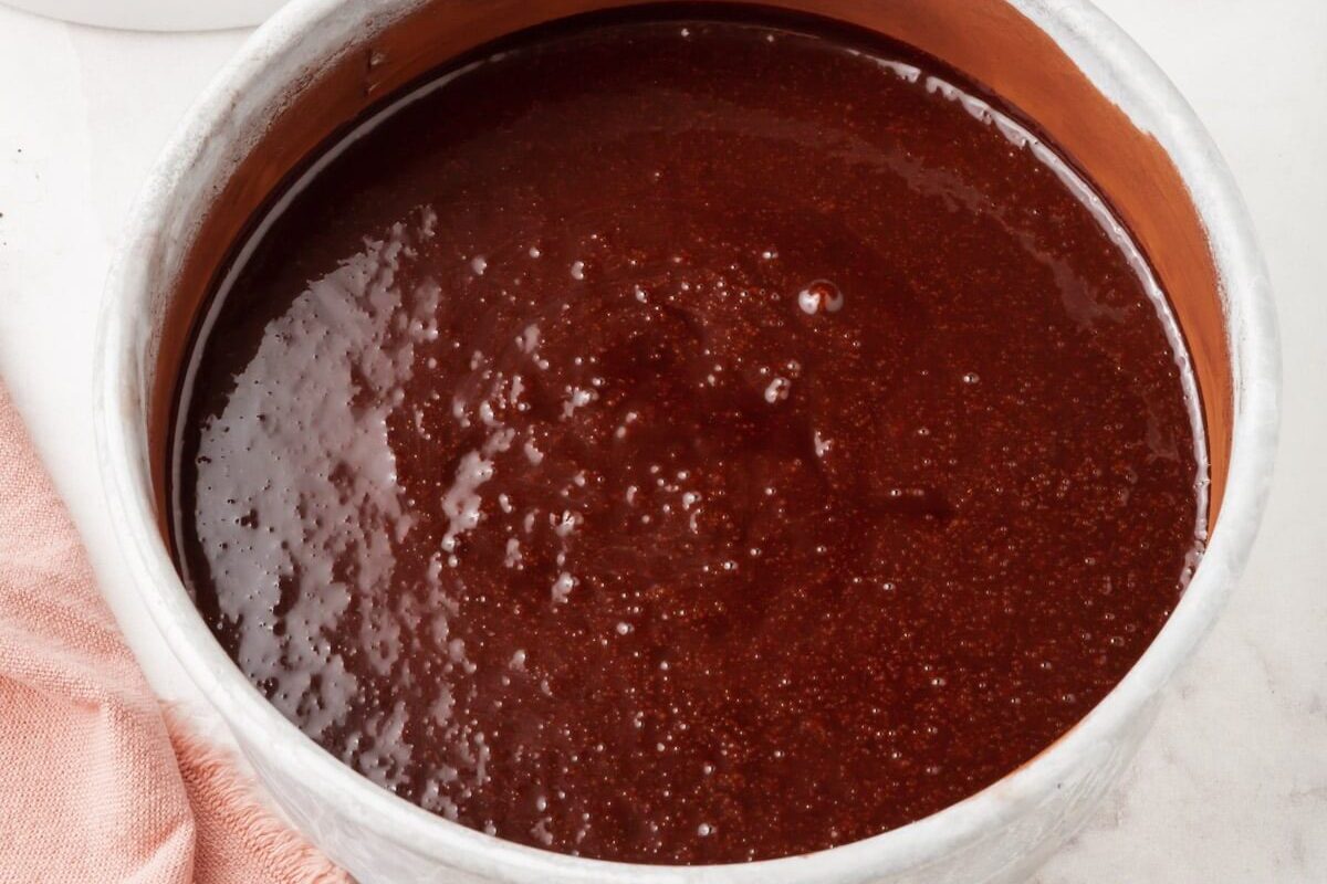 A round cake pan filled with chocolate cake batter sits on a marble countertop next to a pink cloth. Another cake pan is partially visible in the background.