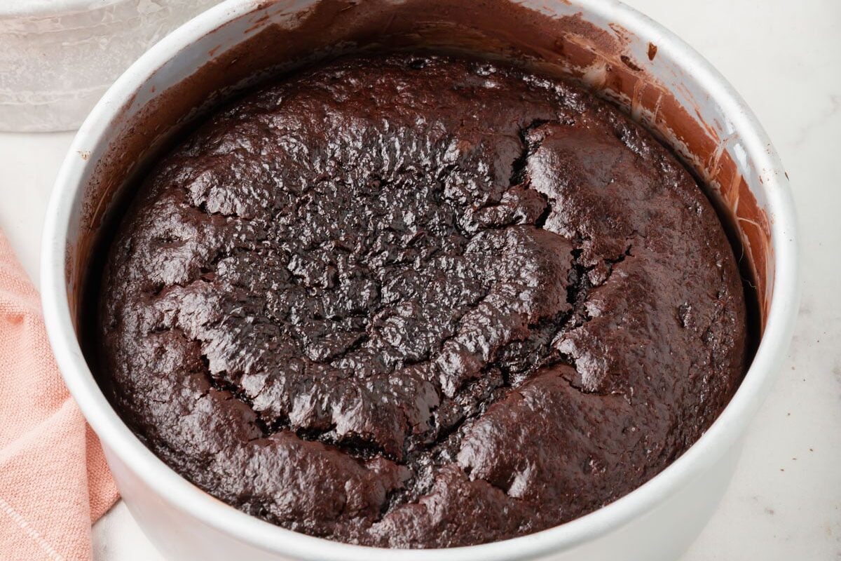 A round, freshly baked chocolate cake in a metal pan sits on a marble surface next to a peach-colored cloth. Another cake pan is partially visible in the background.