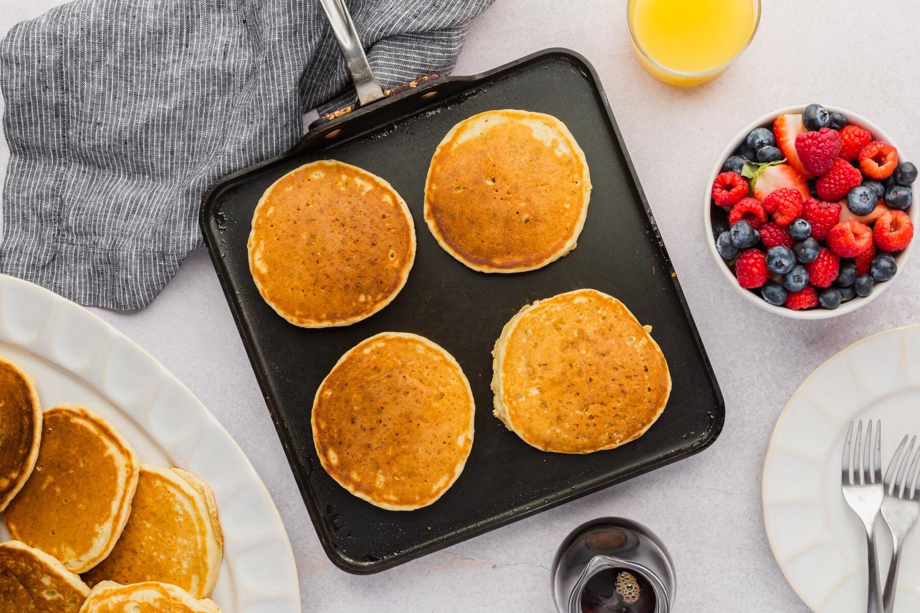 A non-stick griddle with gluten-free panckes on it and a side of maple syrup and fresh berries.