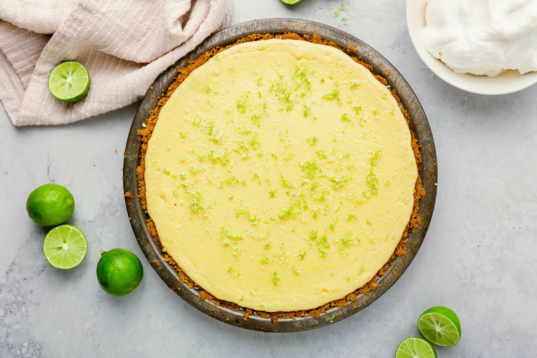 A whole key lime pie with a graham cracker crust, topped with lime zest, sits on a gray surface. Fresh limes, a beige cloth, and a bowl of whipped cream are nearby.