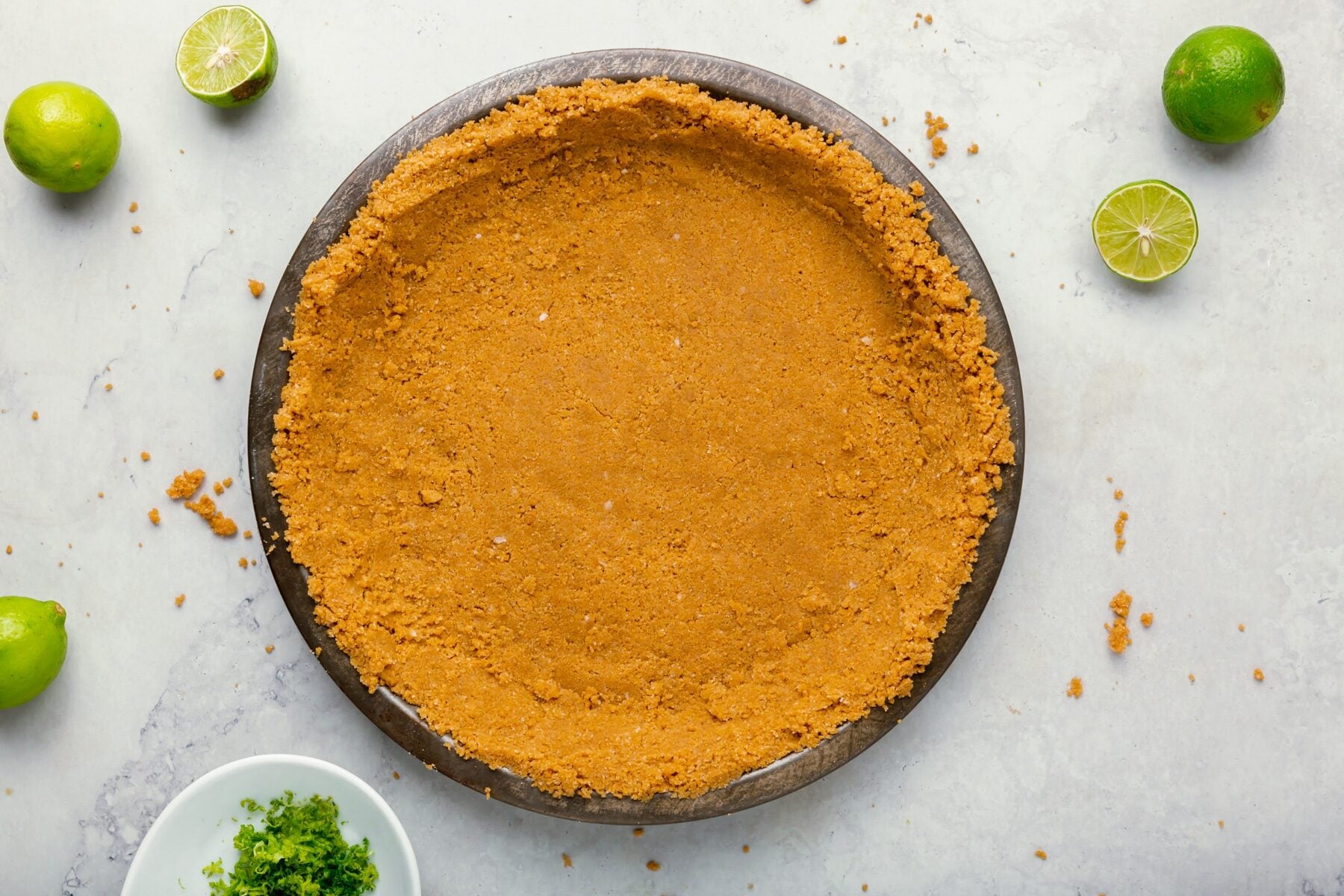 A round pie dish filled with a pressed graham cracker crust sits on a light surface. Whole and halved limes and a small bowl of lime zest are scattered around the dish.