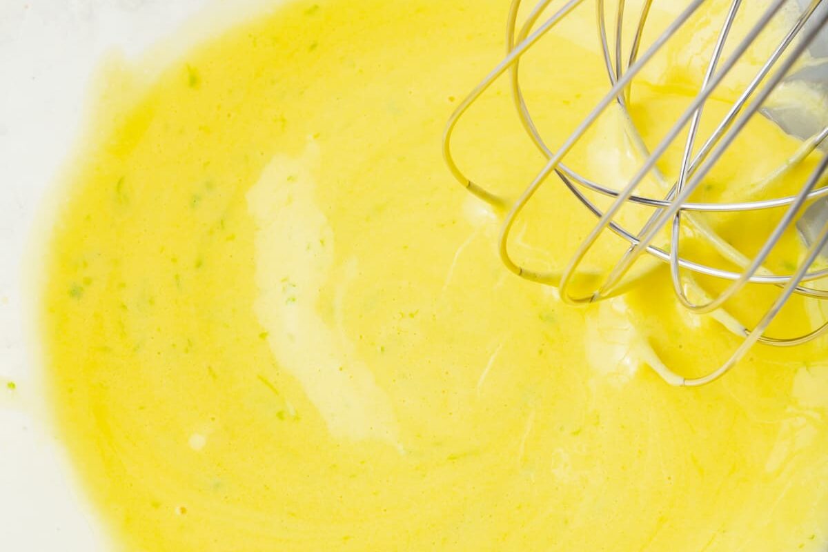 A close-up of a metal whisk mixing a bright yellow batter or sauce in a bowl, with a smooth and creamy texture.