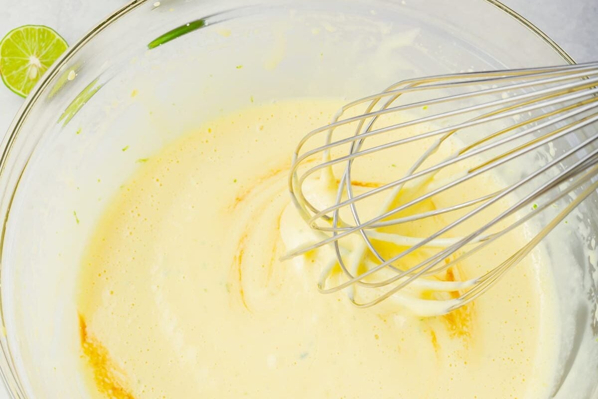 A metal whisk sits in a glass bowl containing a frothy, pale yellow mixture. Bits of green zest and a slice of lime are visible around the edge of the bowl.