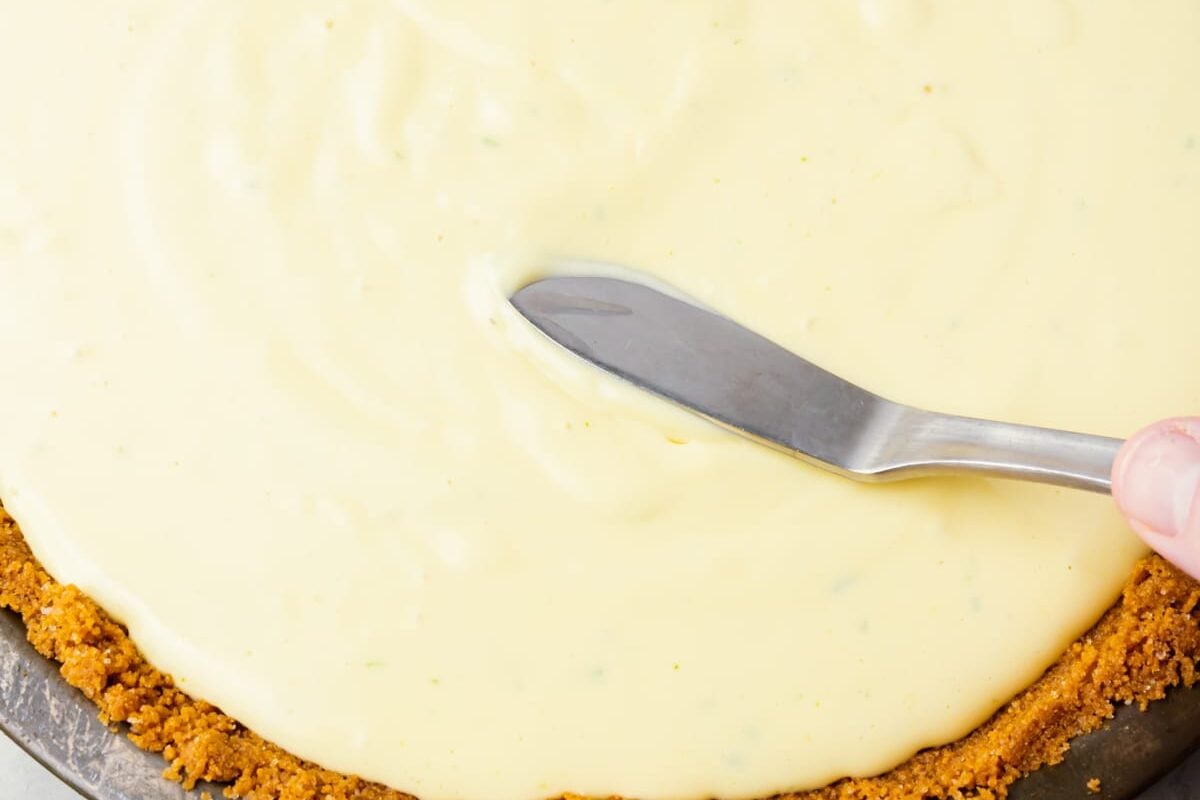A close-up of a hand using a metal spatula to spread creamy key lime pie filling evenly over a gluten-free graham cracker crust in a pie dish.