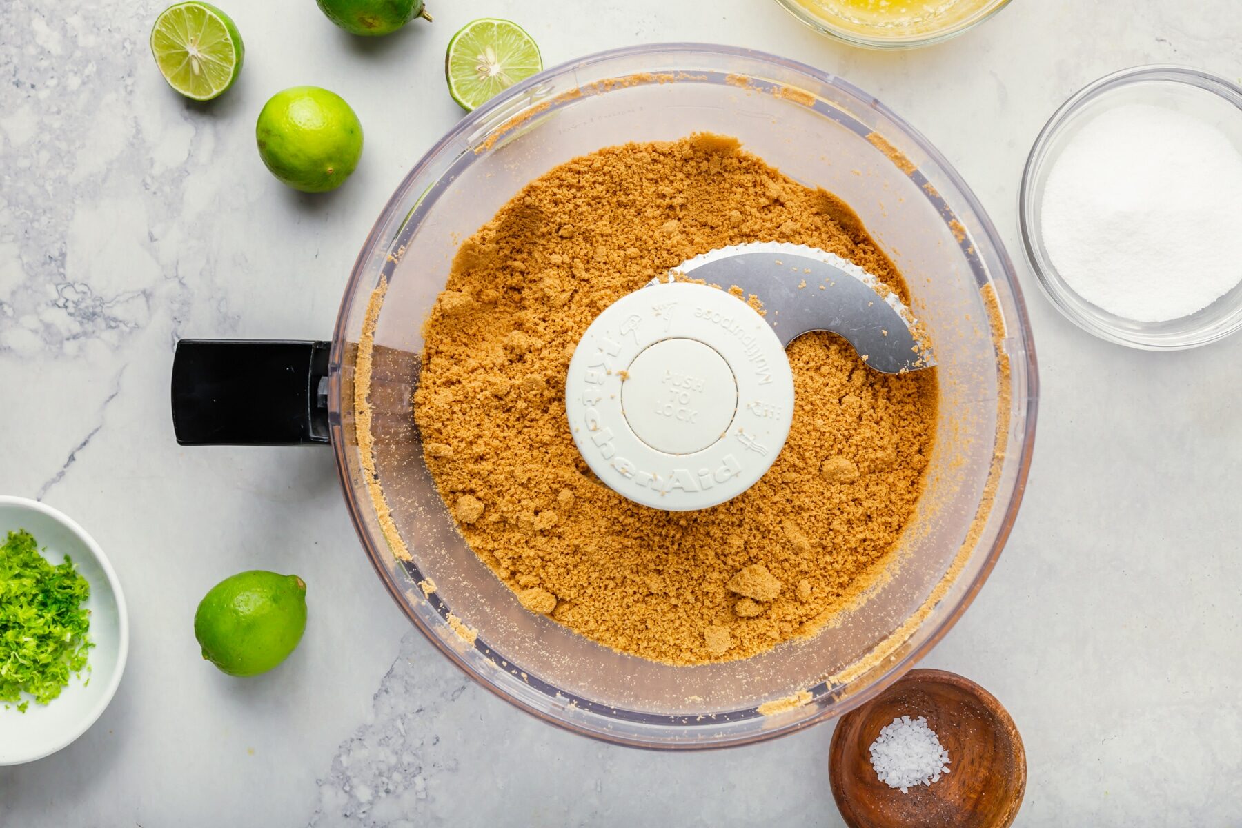 A food processor with crushed graham crackers sits on a marble countertop, surrounded by whole limes, a cut lime, a bowl of sugar, lime zest, salt, and a bowl of liquid ingredients.