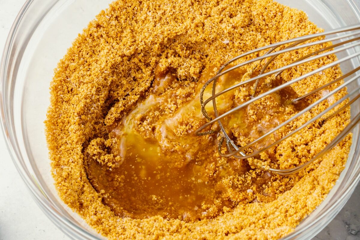 A close-up of a glass bowl with graham cracker crumbs being mixed with melted butter using a metal whisk, preparing a crust mixture.