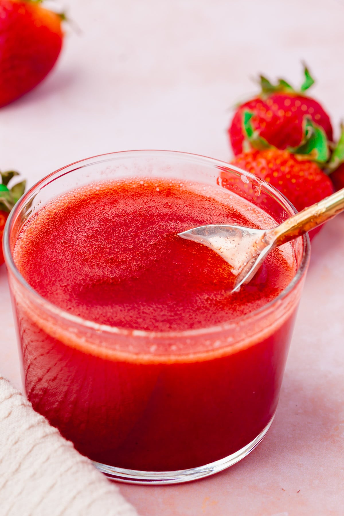 A bowl of strawberry coulis with a spoon inside surrounded by whole strawberries.