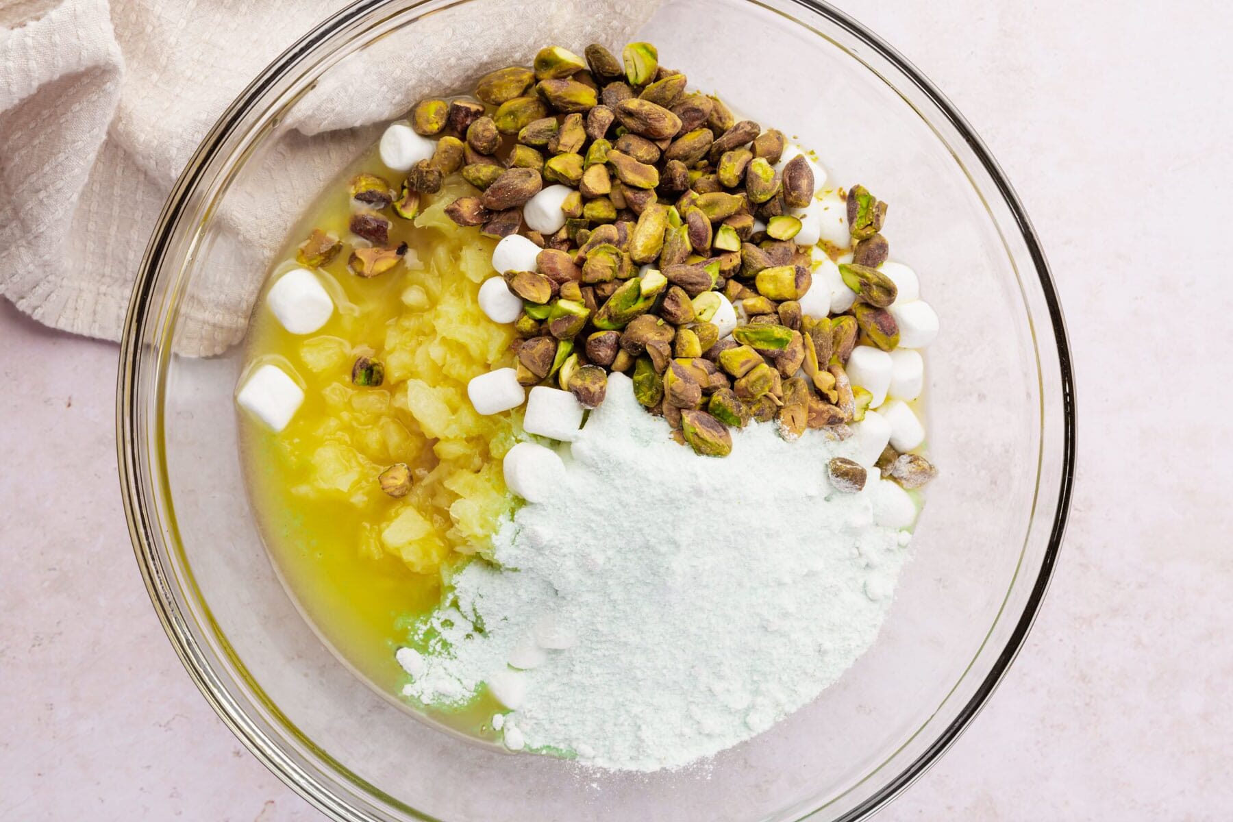 Crushed pineapple, pistachios, marshmallows and pistachio pudding mix in a glass mixing bowl.