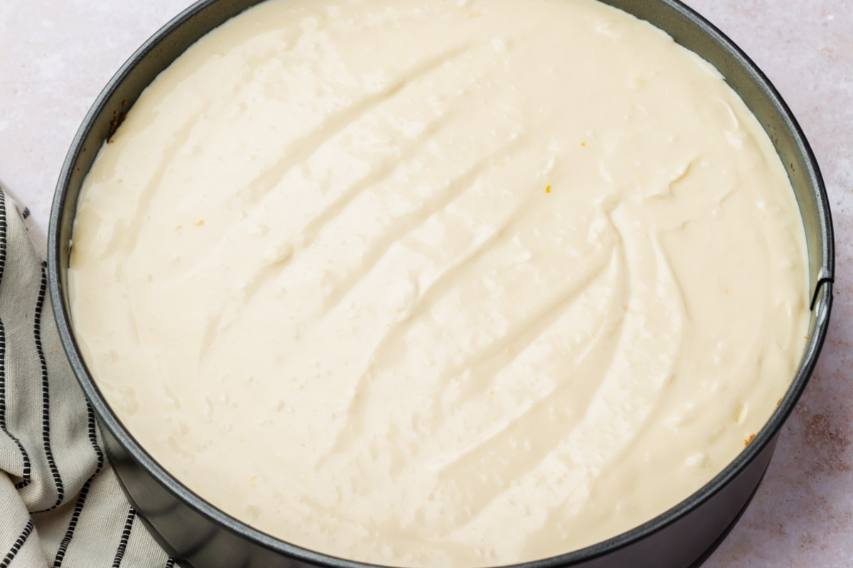 A close-up of gluten free cheesecake batter spread smoothly in a round springform pan, with a striped kitchen towel partially visible on the side.