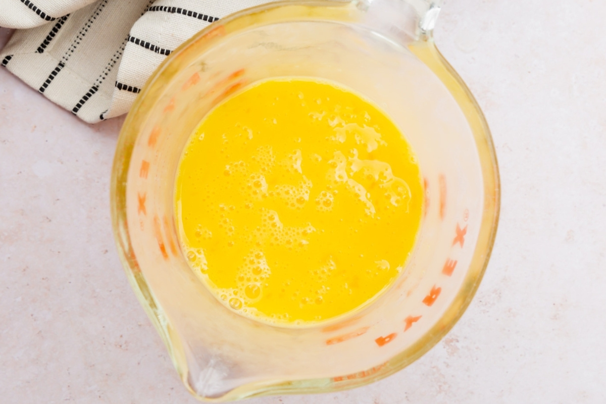 A glass measuring cup filled with whisked eggs sits on a light countertop, ready to help create a gluten free cheesecake, with a striped kitchen towel partially visible in the corner.