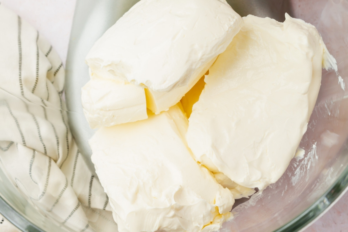 A close-up of several large blocks of cream cheese in a glass mixing bowl, ready to be blended into a delicious gluten free cheesecake, with a striped kitchen towel partially visible to the side.