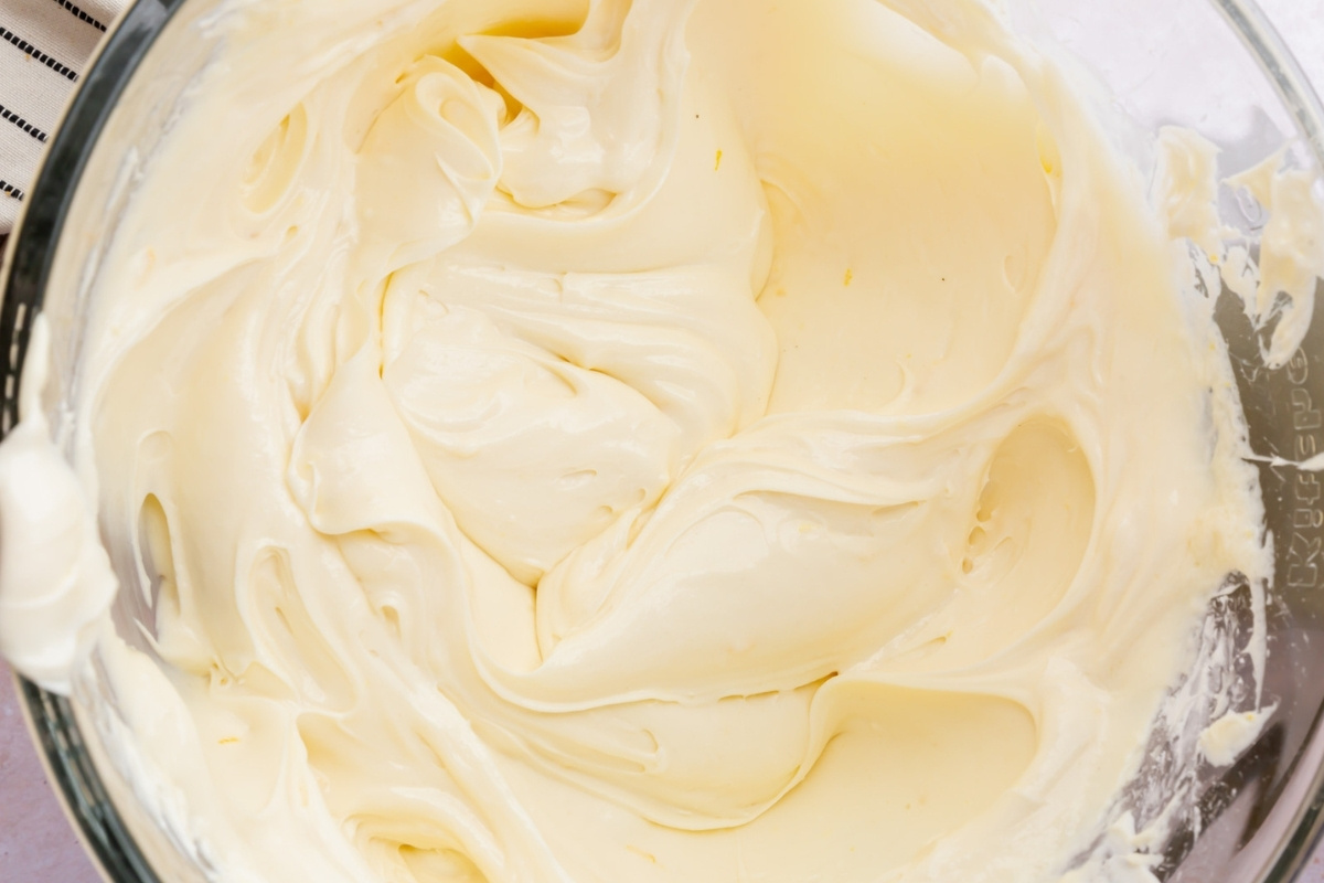 A close-up of smooth, creamy, pale yellow frosting swirled in a glass mixing bowl. The thick, fluffy texture with soft peaks and folds is perfect for topping a gluten free cheesecake.