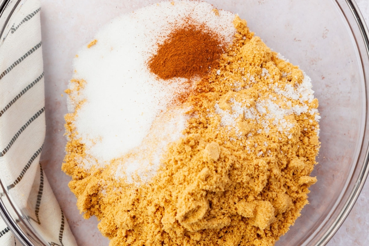 A glass bowl filled with white granulated sugar, light brown sugar, ground cinnamon, and a heap of graham cracker crumbs for a gluten free cheesecake crust. A striped kitchen towel is partially visible on the side.