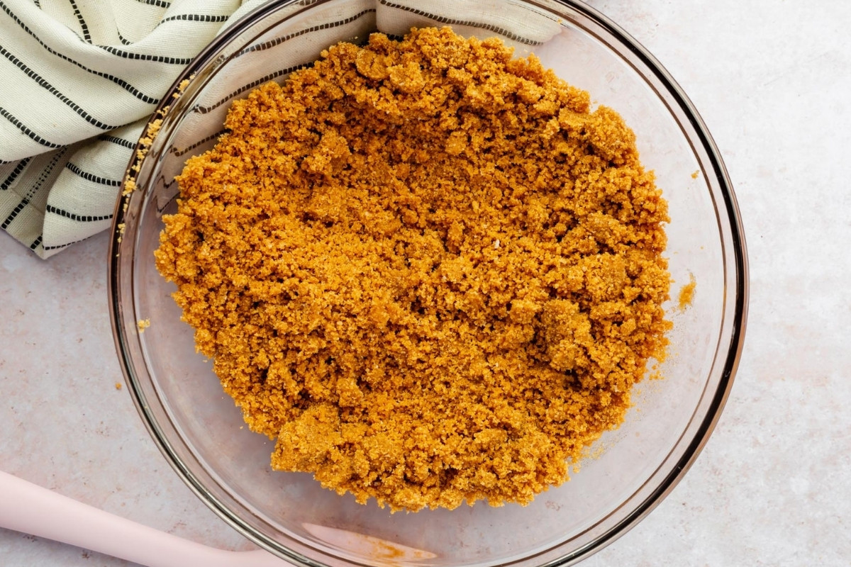 A glass bowl filled with a crumbly graham cracker crust mixture for a gluten free cheesecake sits on a light surface next to a striped kitchen towel.