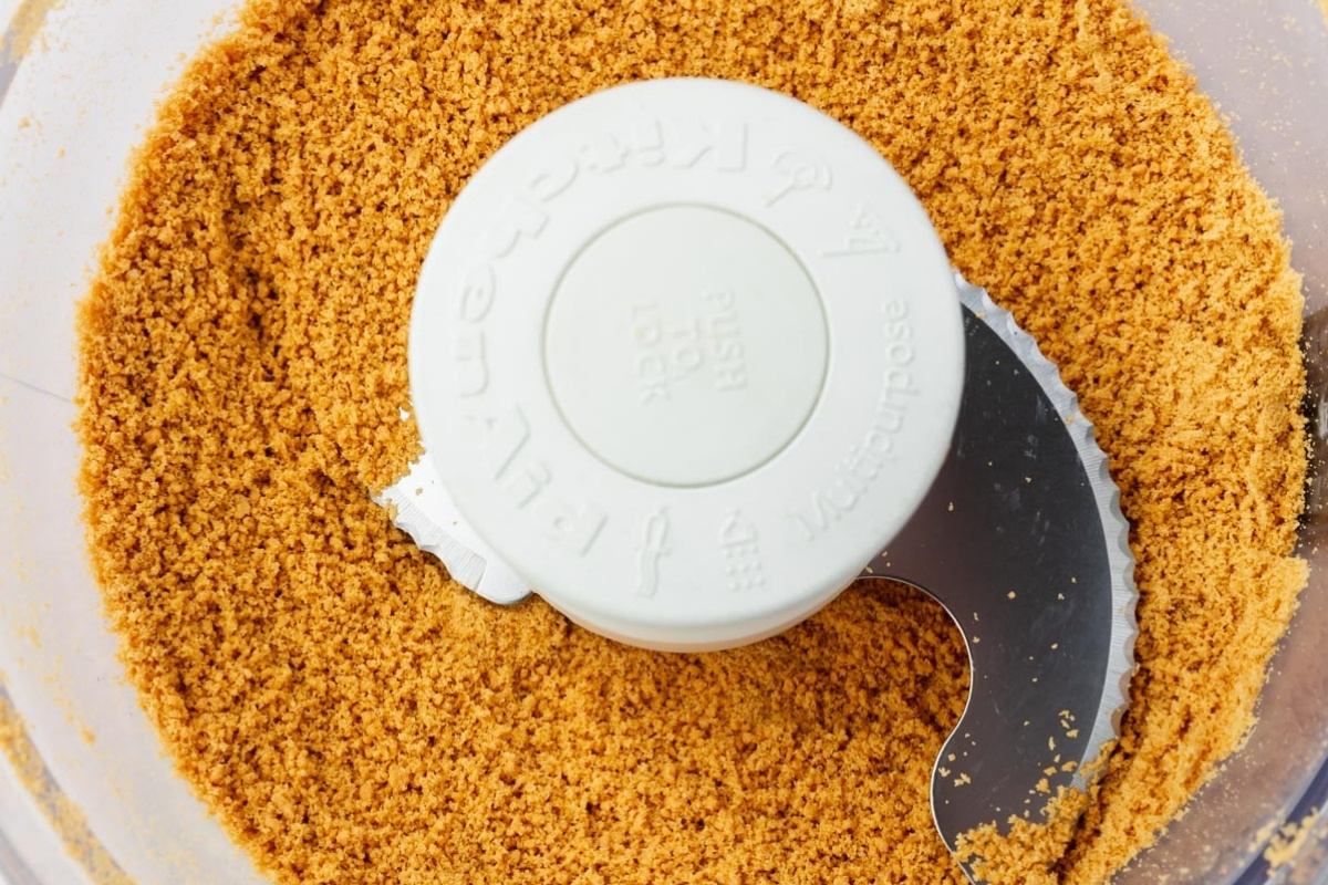Crushed graham crackers in a food processor bowl, surrounding the metal blade and center lid, ready for use in a recipe such as a gluten free cheesecake base or classic pie crust.