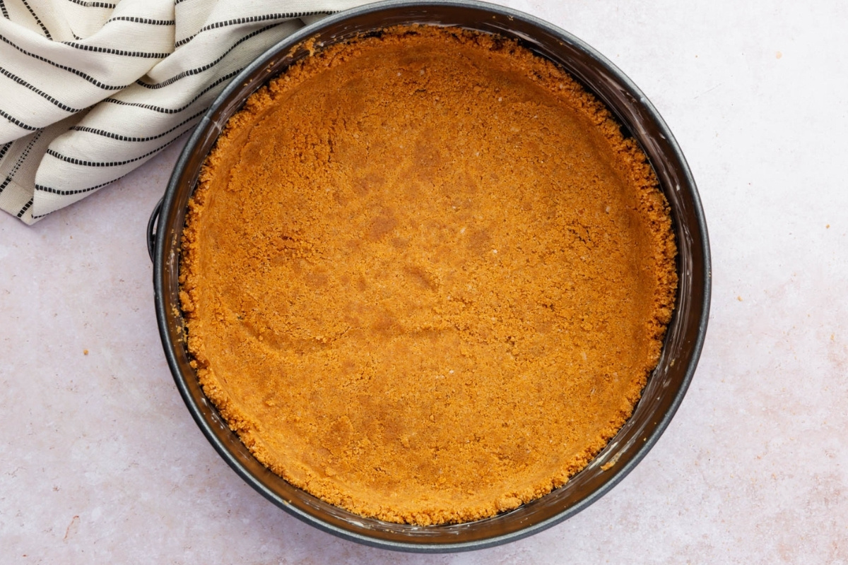 A round baking pan filled with an evenly pressed graham cracker crust for a gluten free cheesecake sits on a light countertop, with a folded white and black striped kitchen towel partially visible in the corner.