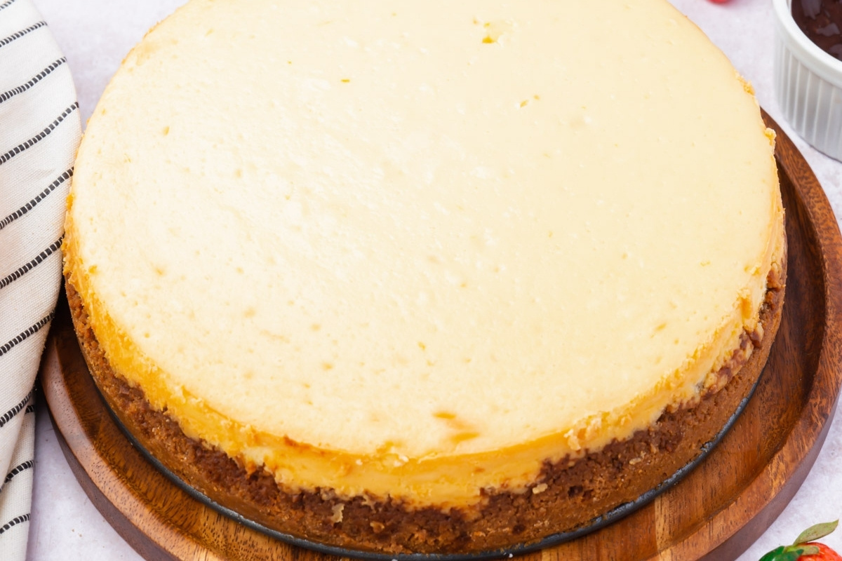 A whole, plain gluten free cheesecake with a golden-brown crust sits on a round wooden board. The cheesecake has a smooth, creamy top with no toppings or decorations. A striped cloth is partially visible on the side.