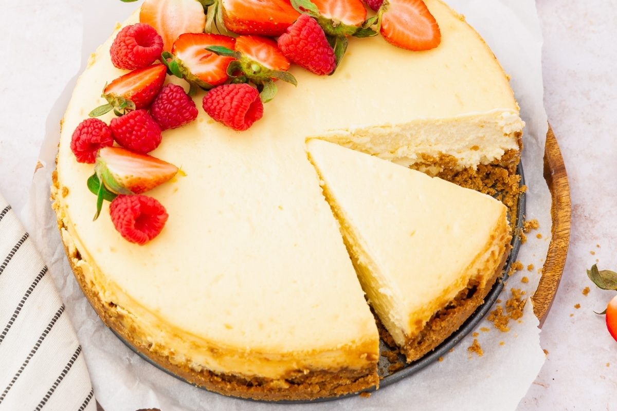 A gluten free cheesecake with a golden crust, topped with fresh strawberries and raspberries. One slice is cut and slightly pulled out, revealing the creamy texture inside. The cake sits on parchment paper on a wooden board.
