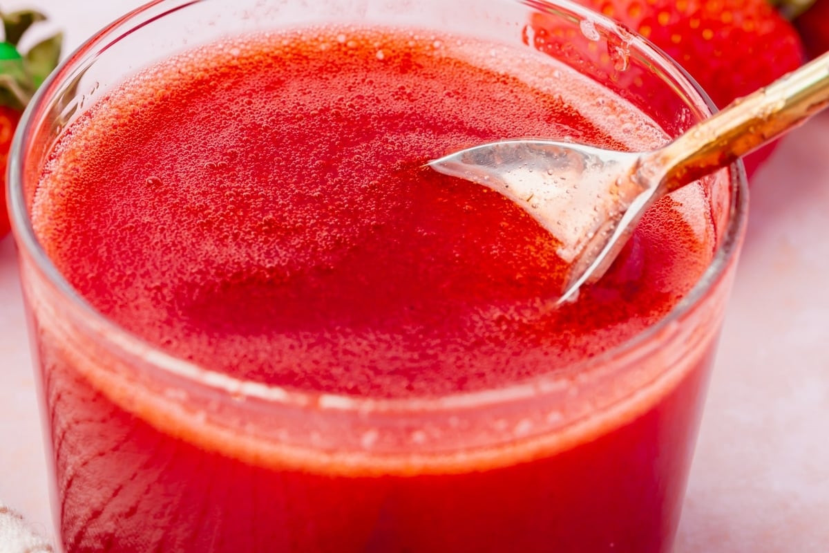 A close-up of a glass filled with bright red strawberry juice, with a spoon resting inside. Fresh strawberries and a hint of strawberry coulis add vibrant color to the background.