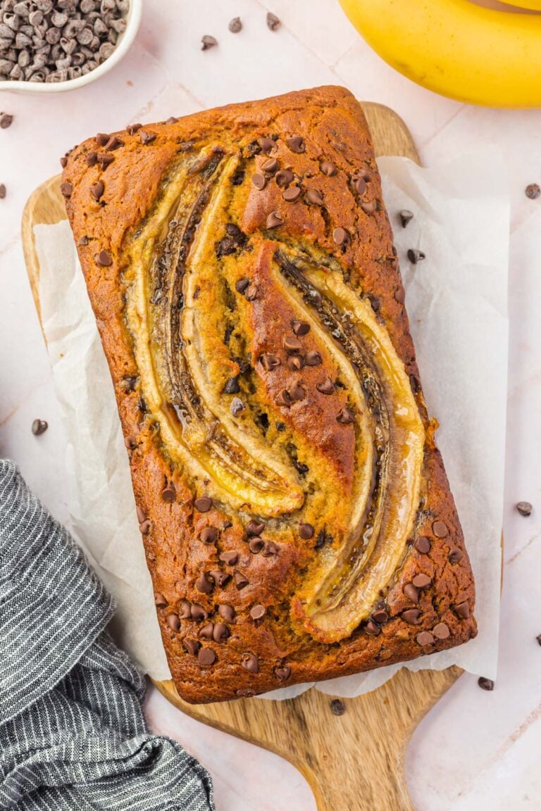 A loaf of gluten-free banana bread on a wooden cutting board next to a banana and a bowl of chocolate chips.