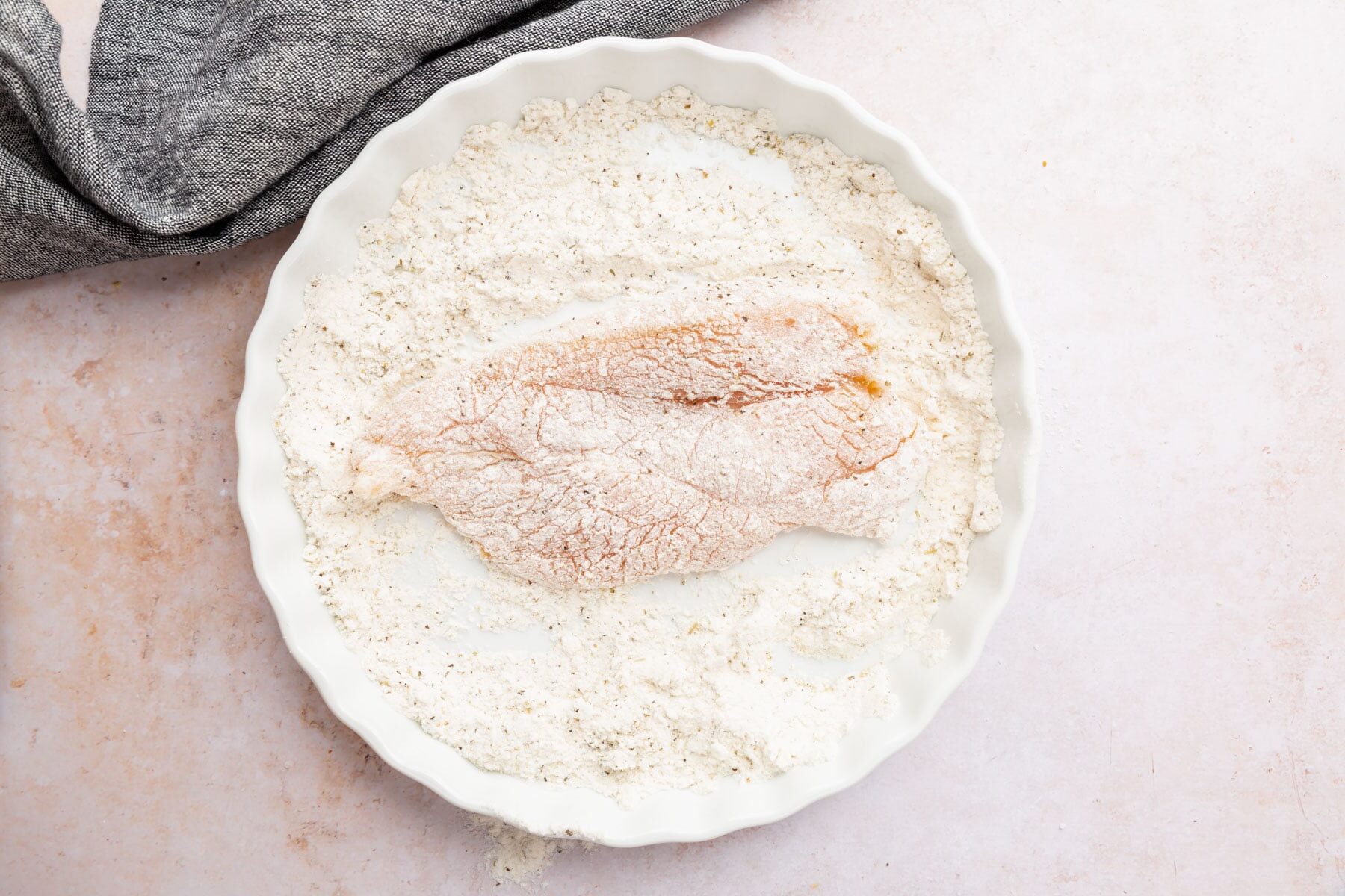 A boneless skinless chicken breast in a bowl of flour mixture coated on one side.