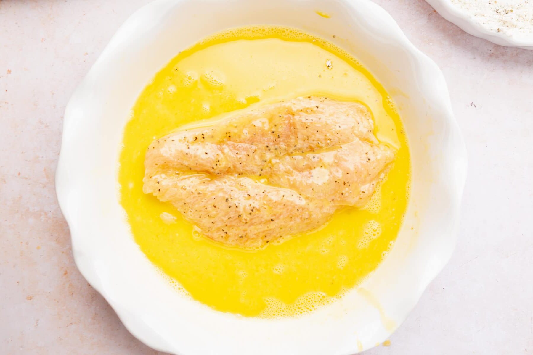 A boneless skinless chicken breast sitting in a bowl of egg mixture.