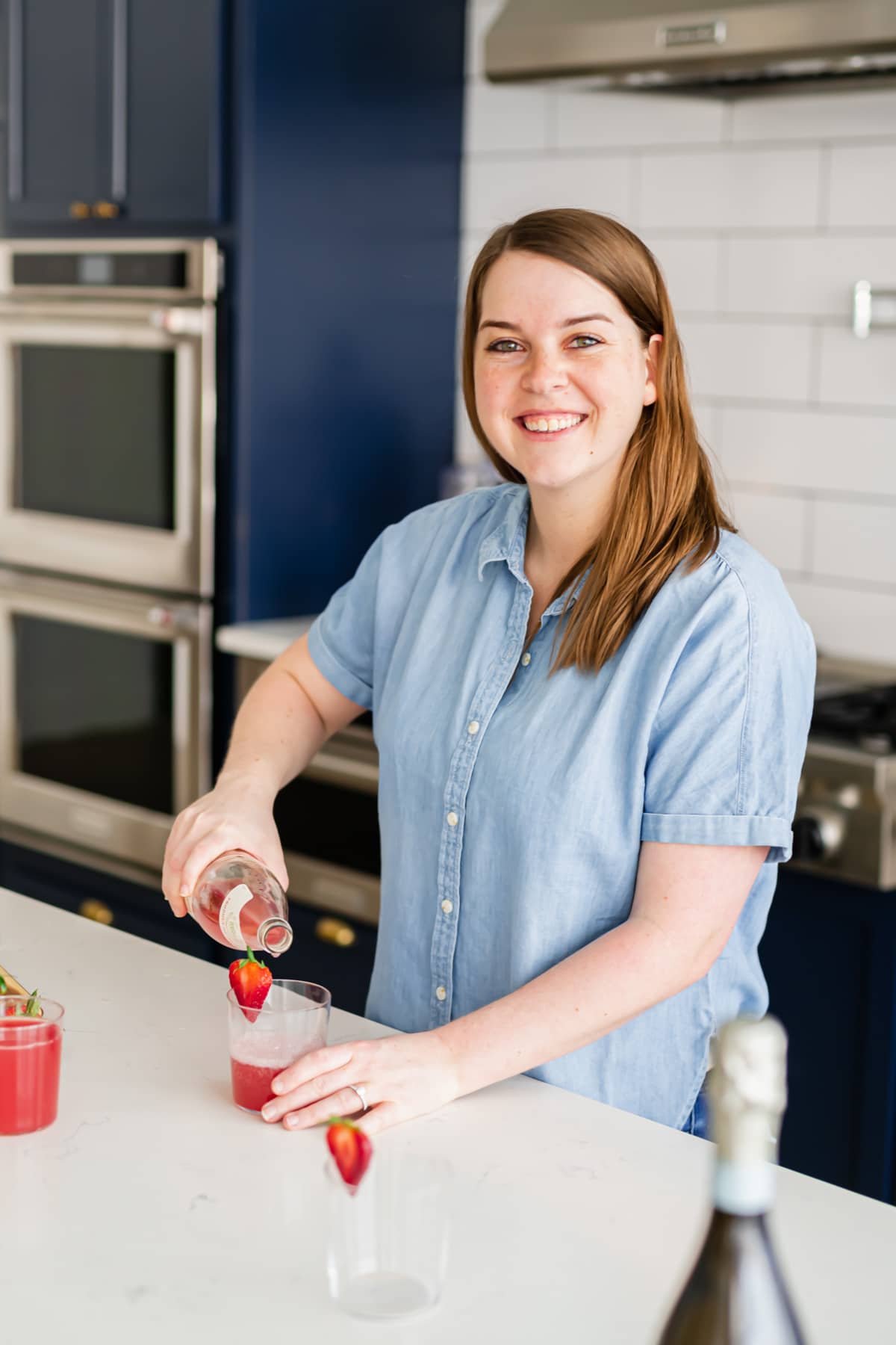 A woman in a blue shirt pouring strawberry cocktail into a glass garnished with a fresh strawberry.