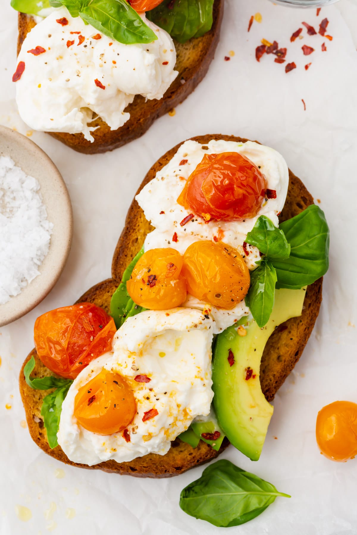 An overhead view of a slice of avocado toast topped with burrata, roasted tomatoes and basil.