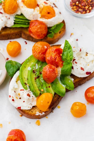 Two slices of avocado toast with burrata and roasted tomatoes on top with a bowl of red pepper flakes.