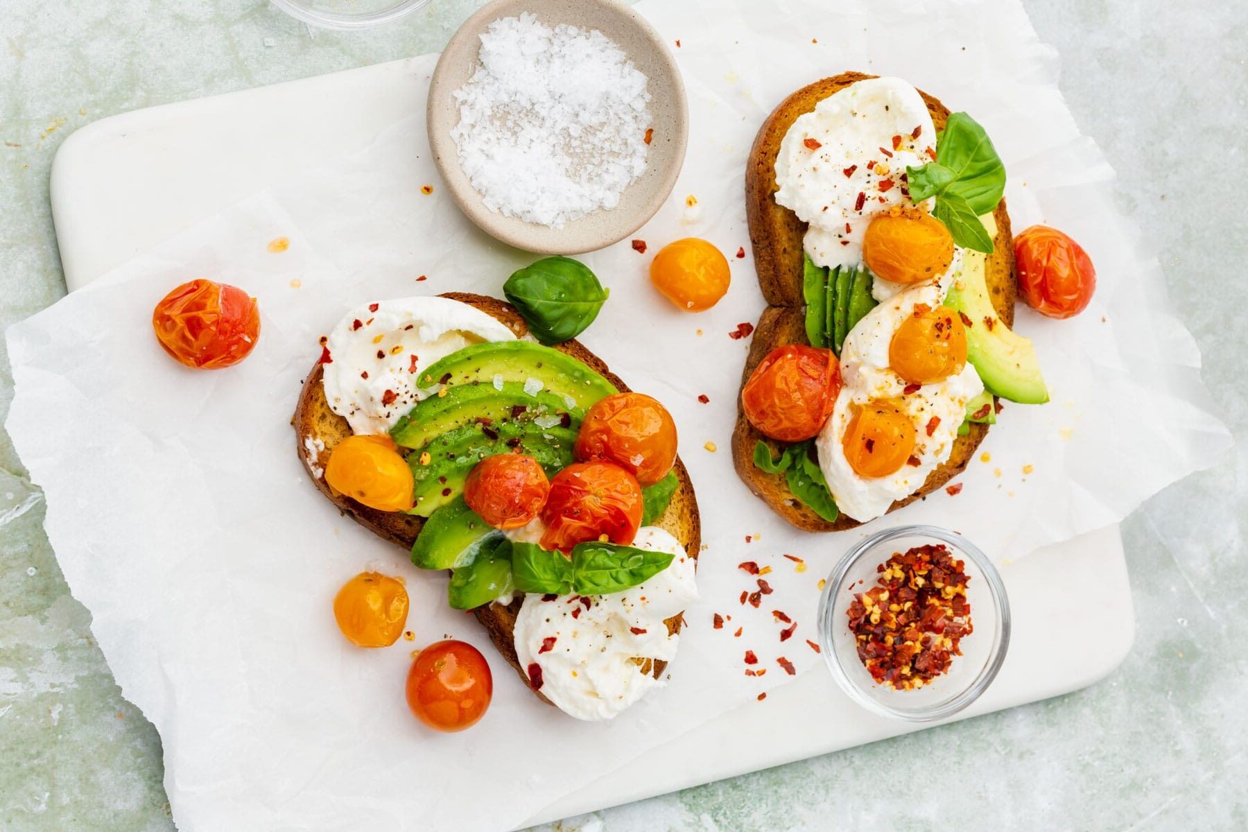 An overhead view of two slices of burrata avocado toast topped with roasted tomatoes with bowls of flaky sea salt and crushed red pepper flakes on the side.