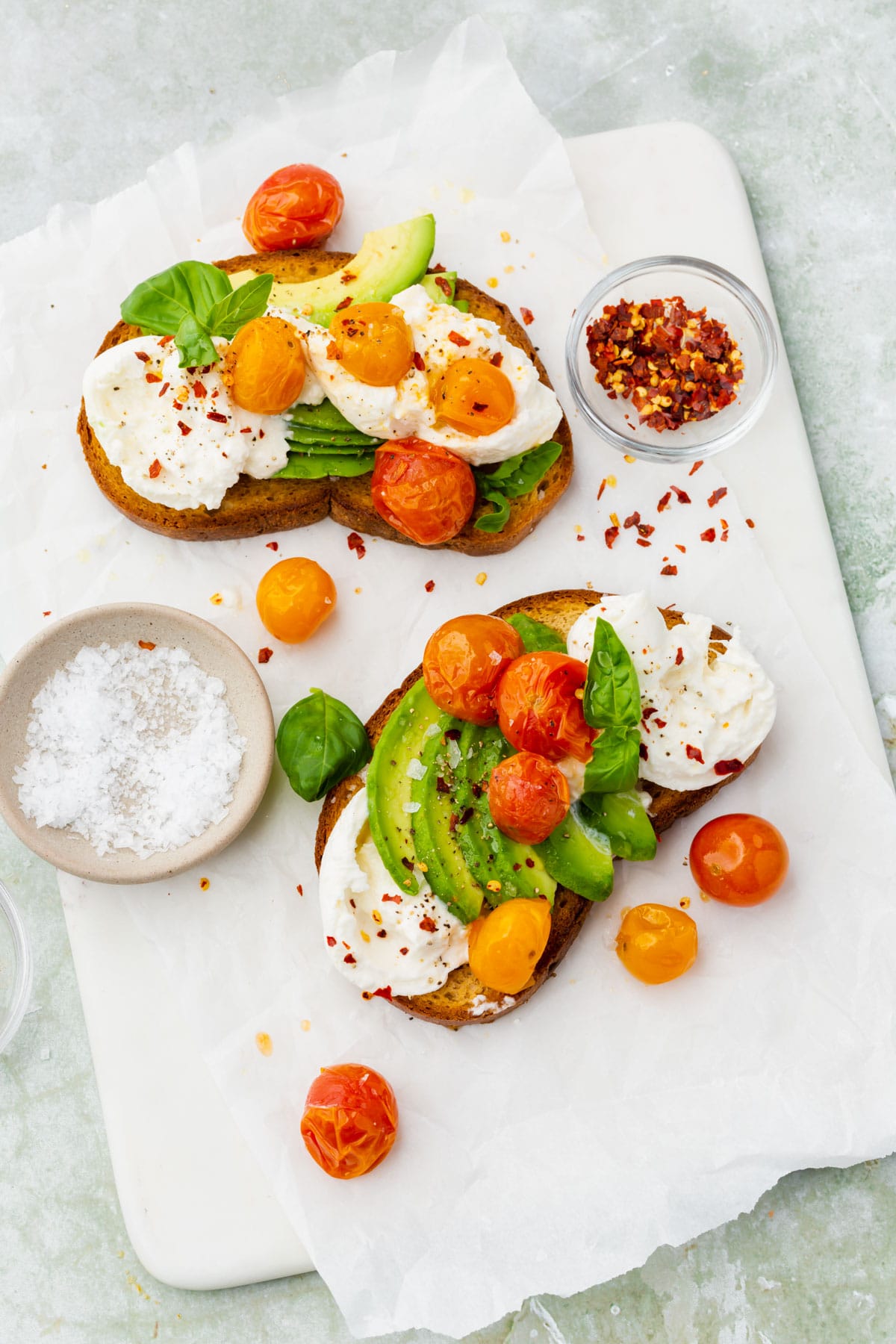 An overhead view of two slices of burrata avocado toast topped with roasted tomatoes with bowls of flaky sea salt and crushed red pepper flakes on the side.