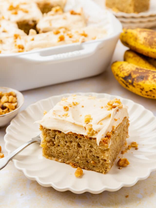 A slice of banana cake on a small plate with bananas and a larger cake in the background.