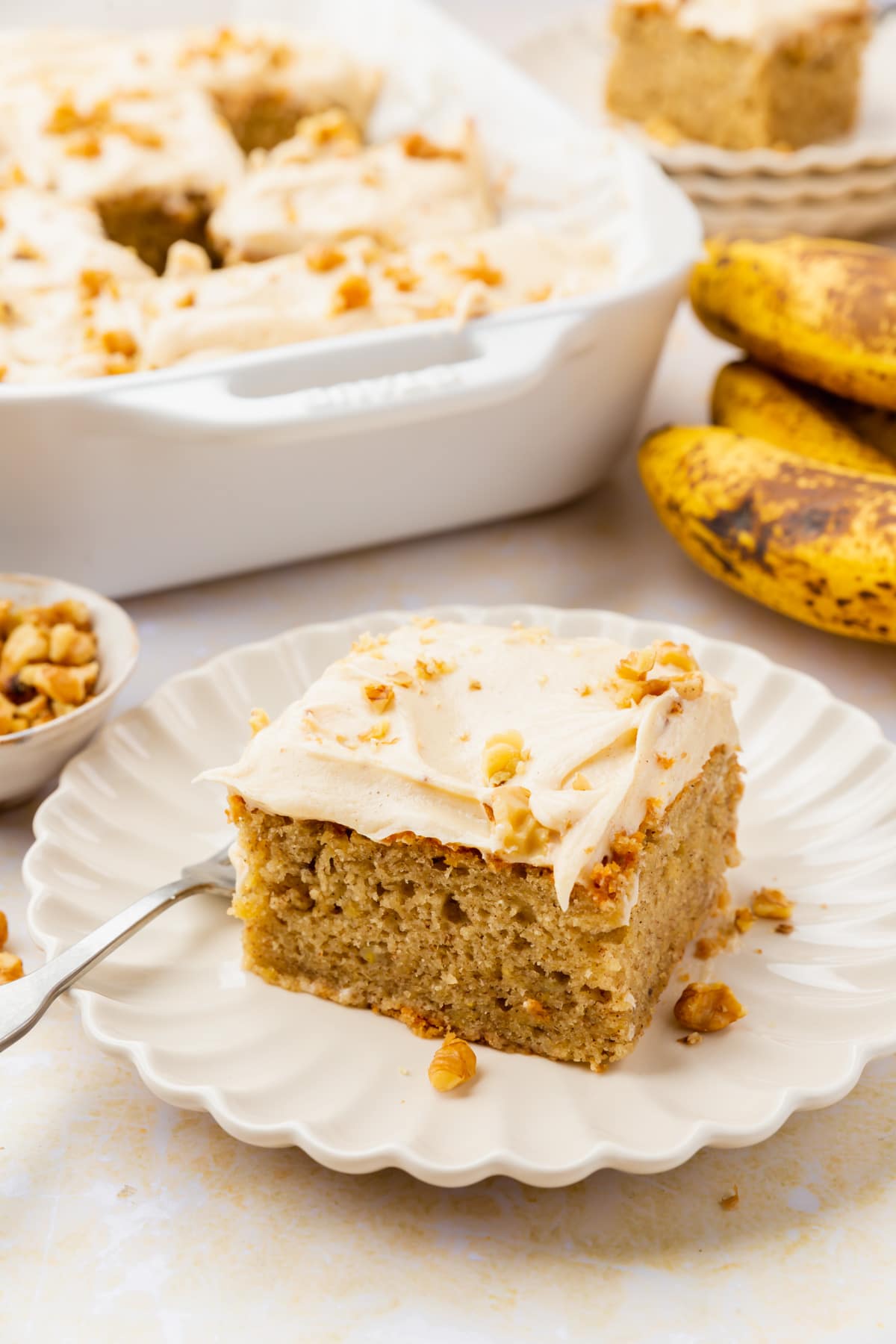 A slice of banana cake on a small plate with bananas and a larger cake in the background. 