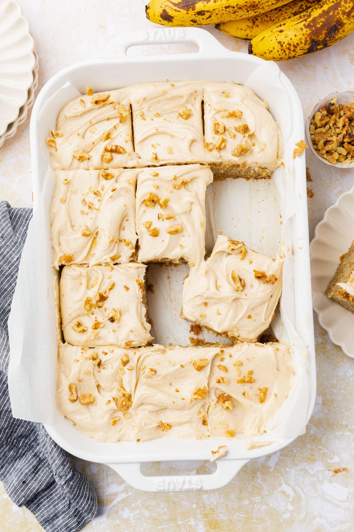 Slices of banana cake in a baking dish topped with cream cheese frosting and walnuts.
