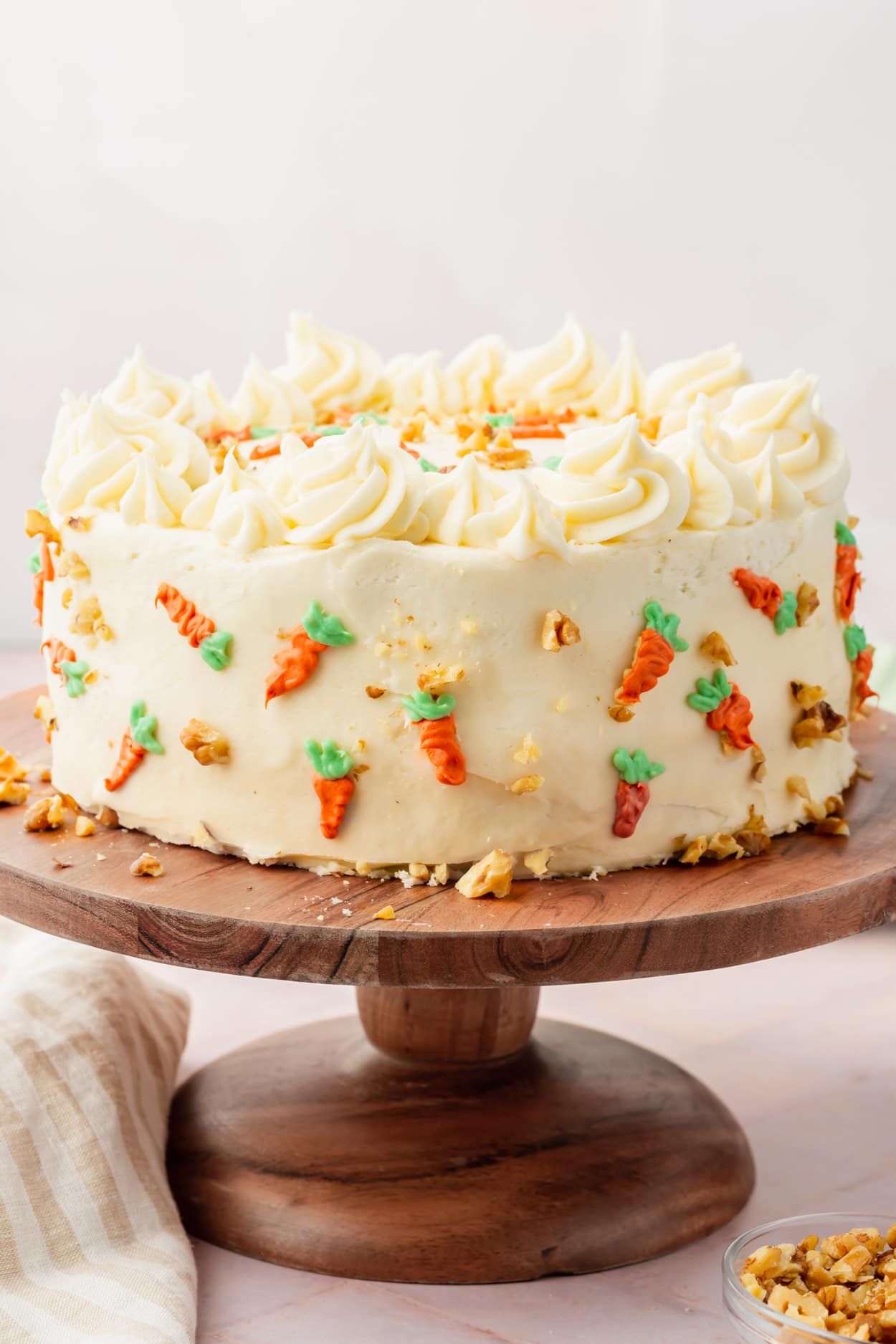 A straight on view of a cake frosted with cream cheese frosting and decorated carrots on a wood cake stand.