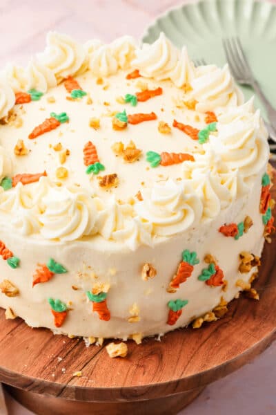 A carrot cake frosted with cream cheese frosting and carrot decorations on a wood cake stand.