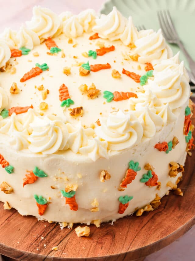 A carrot cake frosted with cream cheese frosting and carrot decorations on a wood cake stand.