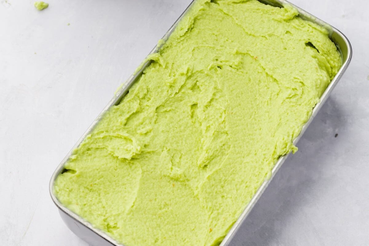 Avocado ice cream in a loaf pan with the ice cream maker bowl in the background.