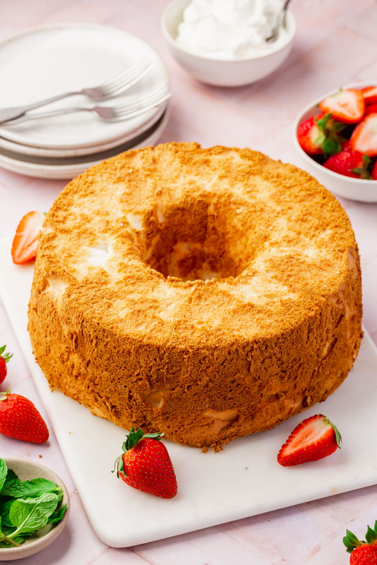 An angel food cake on a serving platter with strawberries scattered around, plates and forks in the background and small bowls of whipped topping, strawberries and mint leaves. 