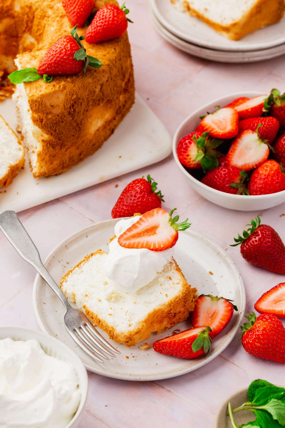 A slice of angel food cake, topped with a dollop of whipped topping and a strawberry, on a dessert plate with a fork and a few sliced strawberries with the angel food cake and a bowl of strawberries in the background. 