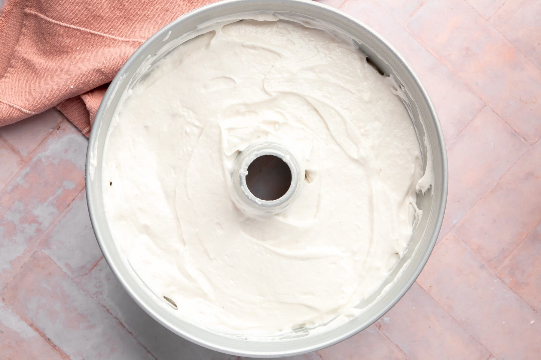 Raw angel food cake batter in a tube pan on top of a pink herringbone table.