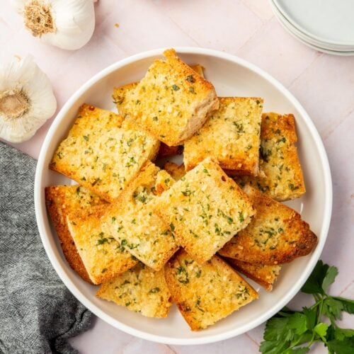 An overhead shot of gluten-free garlic bread in a bowl with garlic bulbs and a bowl of parmesan cheese on the side.