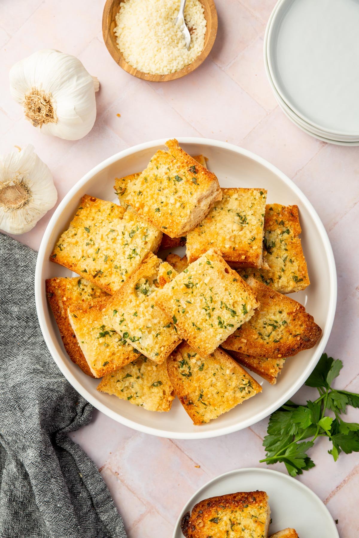 An overhead shot of gluten-free garlic bread in a bowl with garlic bulbs and a bowl of parmesan cheese on the side.