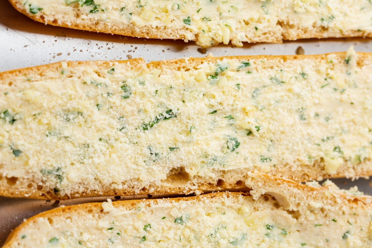 Close-up of sliced bread topped with butter, chopped herbs, minced garlic, and grated cheese, ready to be baked as delicious gluten-free garlic bread.