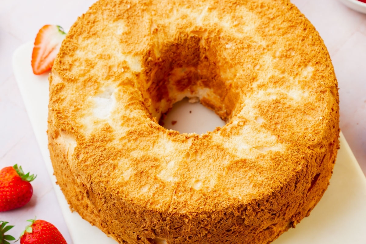 A golden brown Gluten-Free Angel Food Cake with a fluffy texture and a hole in the center, displayed on a white surface with strawberries nearby.