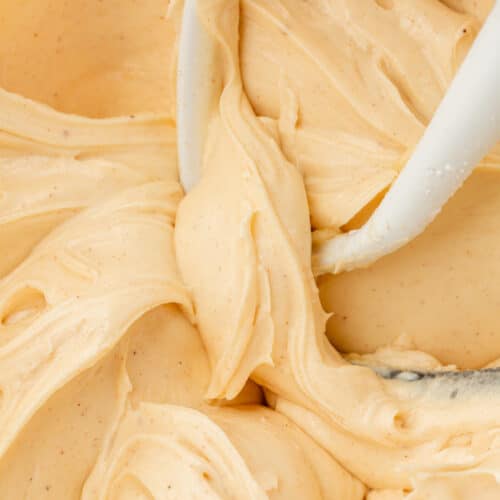 A glass mixing bowl with brown butter cream cheese frosting in it with a paddle attachment from the mixer in the bowl.