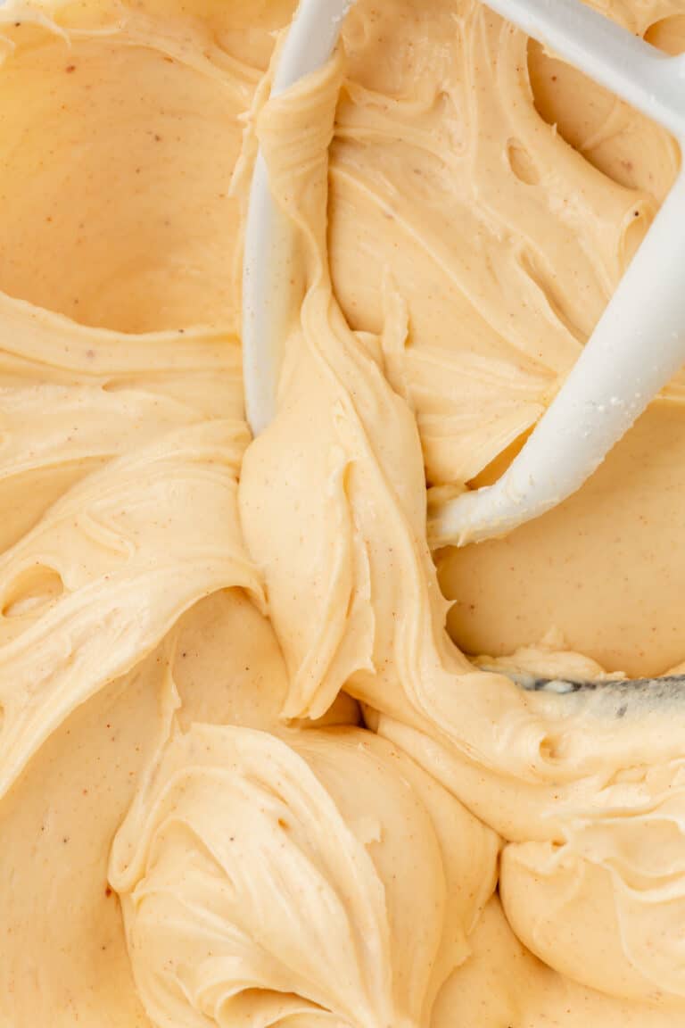 A glass mixing bowl with brown butter cream cheese frosting in it with a paddle attachment from the mixer in the bowl.