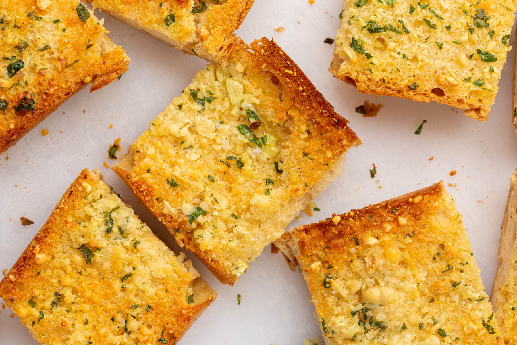 Several slices of golden-brown garlic bread, topped with chopped herbs and small pieces of garlic, are arranged on a white surface. The bread appears crispy and flavorful.